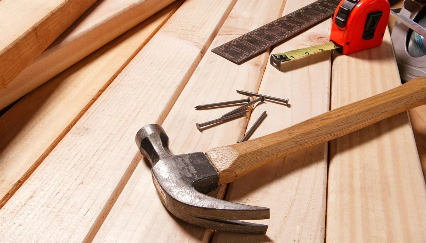 A woodwork setup featuring a hammer, nails, measuring tape, square ruler, and planks of wood.