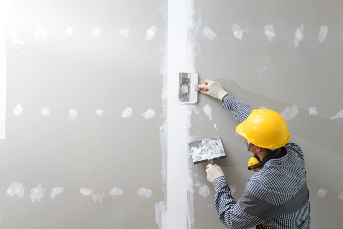 A person wearing a yellow hard hat and gloves applies joint compound on a drywall seam using a taping knife and a mud pan.