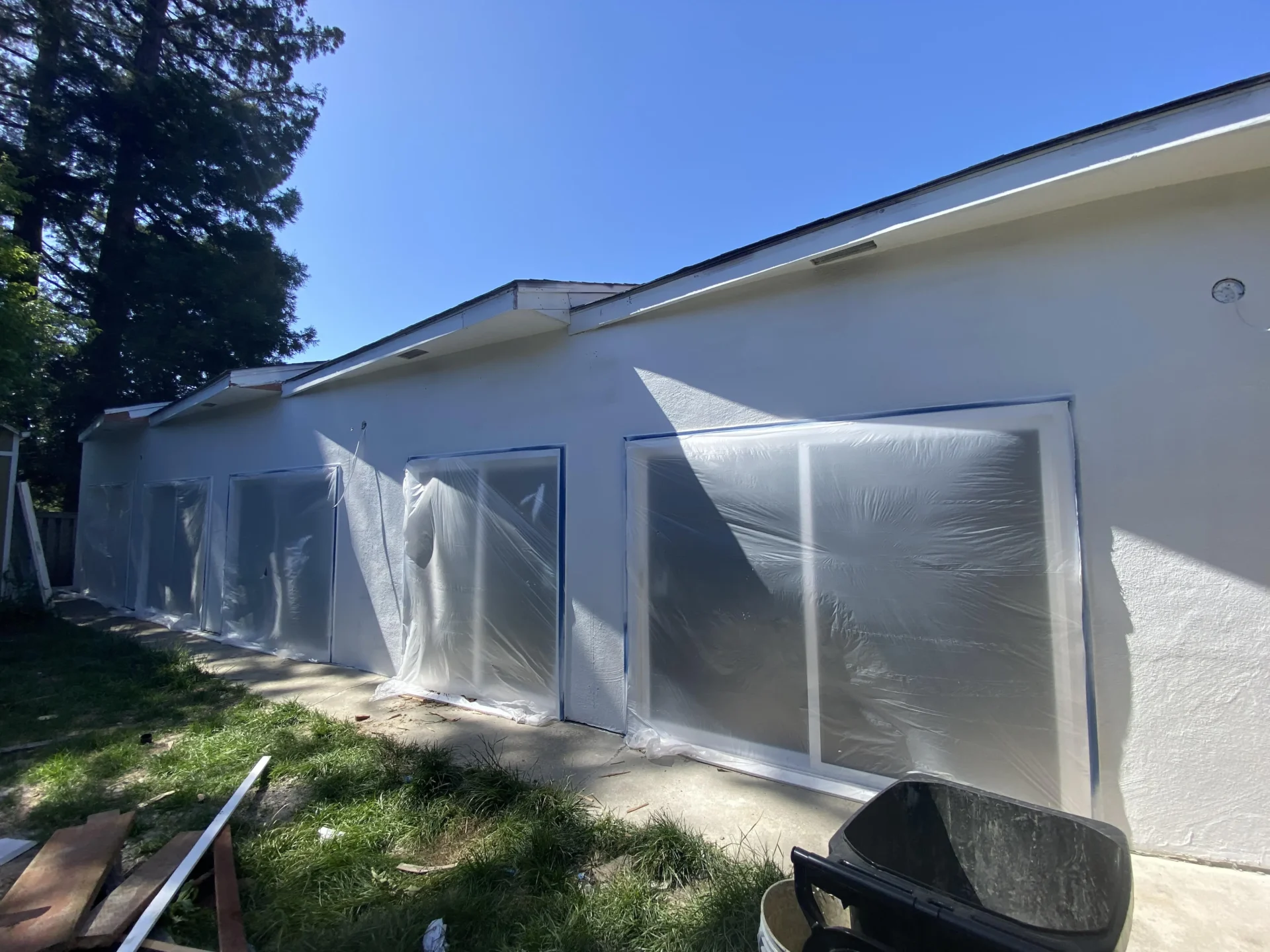 A row of windows on a single-story building covered with plastic sheeting, part of an outdoor renovation project. A black wheelbarrow and some construction materials are in the foreground. exterior painting