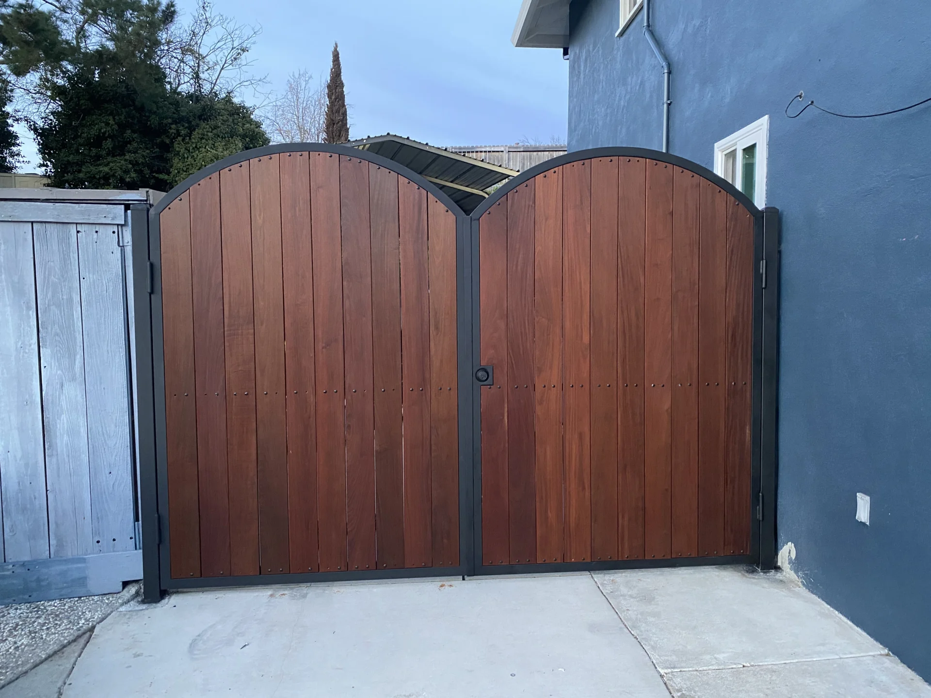 A closed wooden double gate with arched tops and black metal frames is seen attached to a blue house. The gate is flanked by a metal fence on the left and a concrete path below.