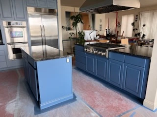 A modern kitchen with blue cabinetry, a large island, stainless steel refrigerator, and a built-in oven. The flooring appears to be under construction.