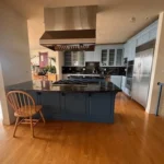 A modern building painters kitchen with a central island, black granite countertops, blue cabinetry, stainless steel appliances, and a wooden chair. The floor is wooden and there's a window in the background.