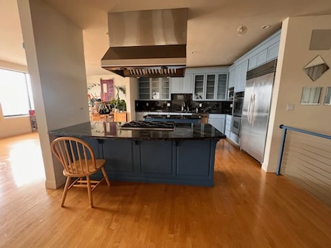 A modern building painters kitchen with a central island, black granite countertops, blue cabinetry, stainless steel appliances, and a wooden chair. The floor is wooden and there's a window in the background.